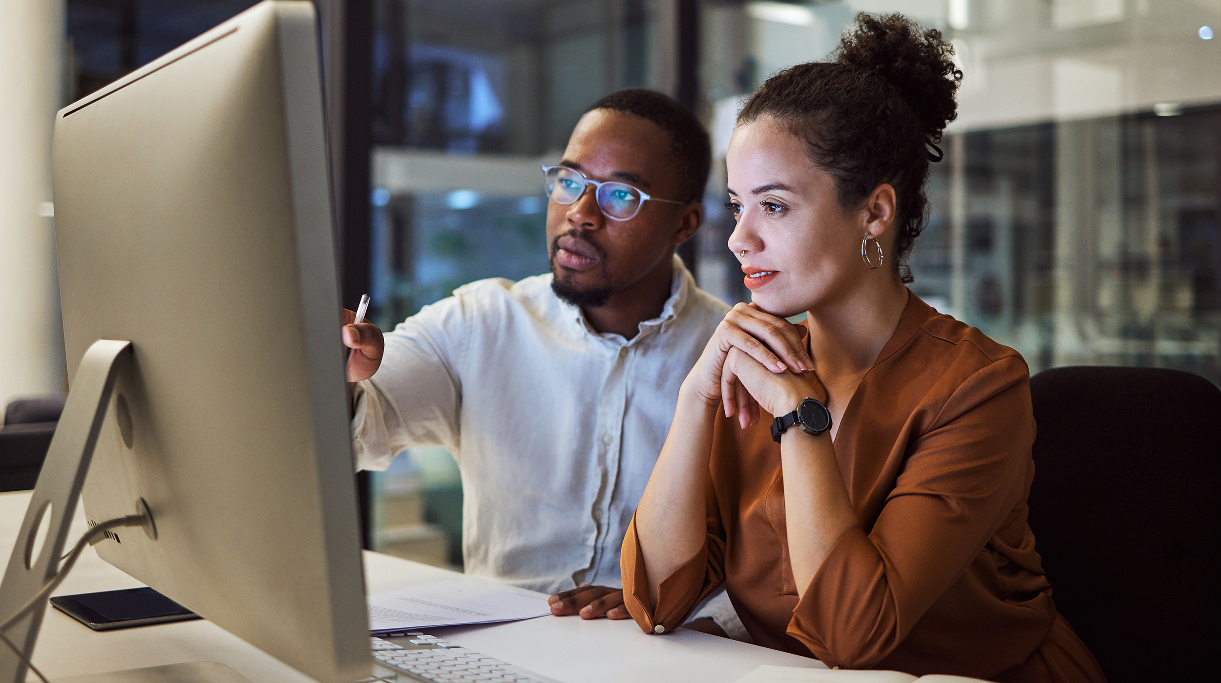 A man and woman sit together at a desk and work together on a desktop computer